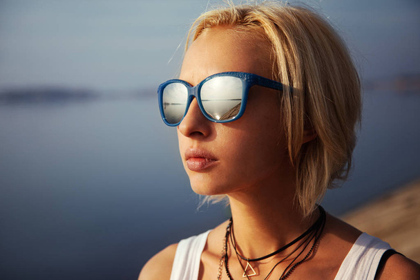 stylová žena s krátkými vlasy stojící v slunečních brýlích v blízkosti řeky ve městě v létě - Fotografie, Obrázek