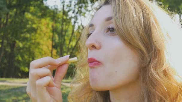 Close up outdoors portrait of young blonde woman eating french fries sitting on the bench in the park. Unhealthy eating, lunch time concepts - Imágenes, Vídeo