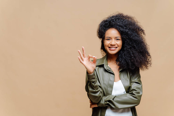 Indoor shot of pleasant looking curly woman has pleasant smile, makes okay gesture, excellent sign, gives approval, dressed in fashionable leather shirt, isolated over brown wall, blank space on left - Photo, Image