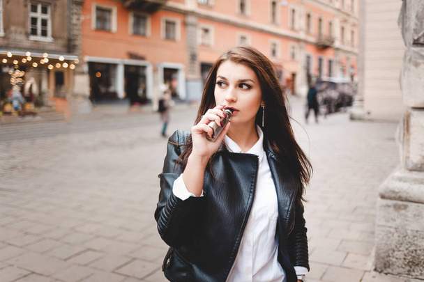 Strong girl in a black leather jacket smokes an electronic cigarette on a city street - Foto, Bild