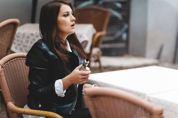 Pretty lady holding electronic cigarette in her hand while sitting at a cafe table - Foto, Imagen