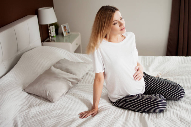 Calm pregnant happy woman is sitting on a light bed with pillows, legs crossed, resting, thinking. - Photo, image