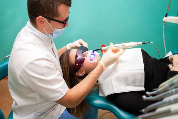 Οδοντίατρος χρησιμοποιώντας λεύκανση υπεριώδους λαμπτήρα στα δόντια της. Νεαρή ασθενής στην οδοντιατρική κλινική βρίσκεται σε γυαλιά UV στην καρέκλα του οδοντιάτρου - Φωτογραφία, εικόνα