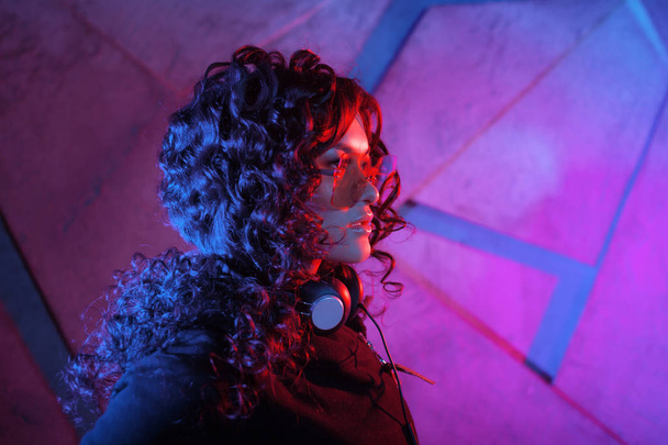 Portrait of young curly pensive sensual woman in big headphones, removed from the head, and shades, on neon background. - Foto, Bild