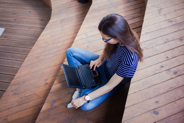 A brunette girl, a student with long hair, with glasses sitting cross-legged on wooden steps and typing on laptop. - Photo, Image