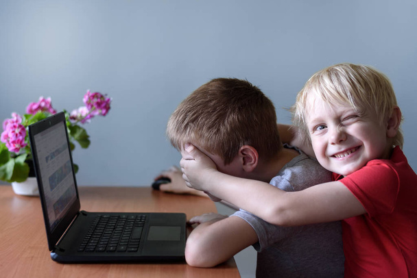 笑顔の少年はノートパソコンを使う友人に彼の目に彼の手を置きます.危険な内容だ子供と道具. - 写真・画像