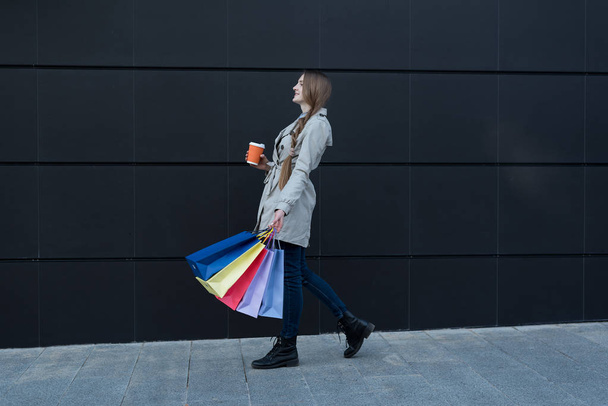 Szczęśliwa młoda kobieta z kolorowymi torbami i papierową filiżanką spacerującą po ulicy. Czarna ściana na tle - Zdjęcie, obraz