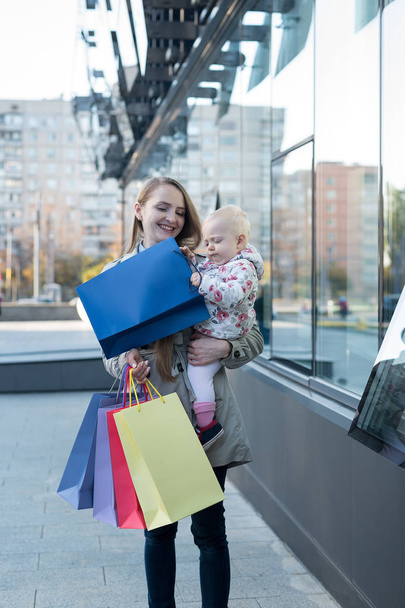 Szczęśliwa młoda matka z córeczką w ramionach i zabawą z torbami. Dzień zakupów. Centrum handlowe na tle - Zdjęcie, obraz