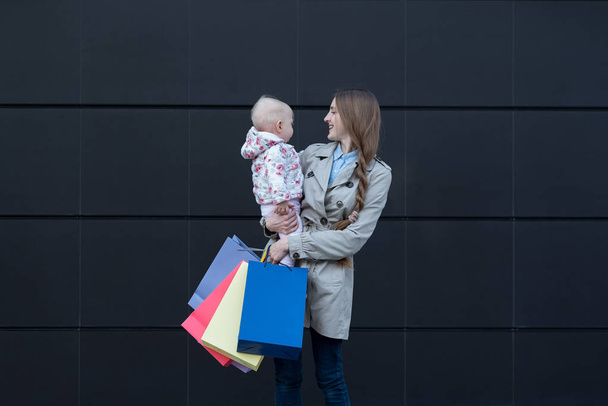 Young mother with little daughter on the arms and shopping bags in hand. Black street wall on background - Foto, imagen