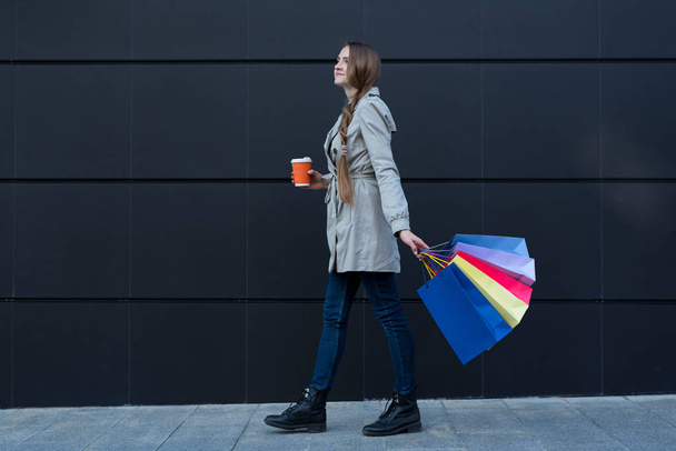 Щаслива молода жінка з різнокольоровими сумками і паперовою чашкою, що йде по вулиці. Чорна стіна на фоні
 - Фото, зображення