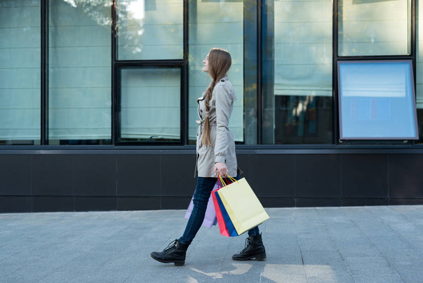Jonge vrouw met kleurrijke tassen die op straat lopen. Mall op achtergrond - Foto, afbeelding