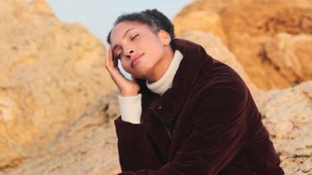 Όμορφη τρυφερή Αφρο-Αμερικανίδα με κατεβασμένο σακάκι που ποζάρει ονειρεμένα πάνω σε πέτρες δίπλα στη θάλασσα - Πλάνα, βίντεο