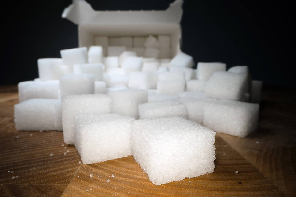 Macro shot of sugar cubes next to carton box on wooden table. Unhealthy food sweetener, sweet crystal cubes - Photo, Image
