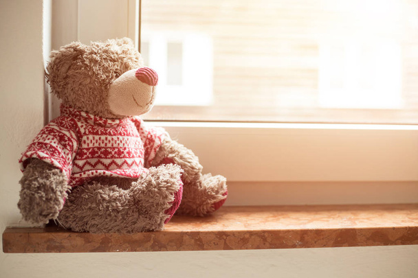 Cute teddy bear is sitting on the windowsill, looking out of the window - Фото, изображение