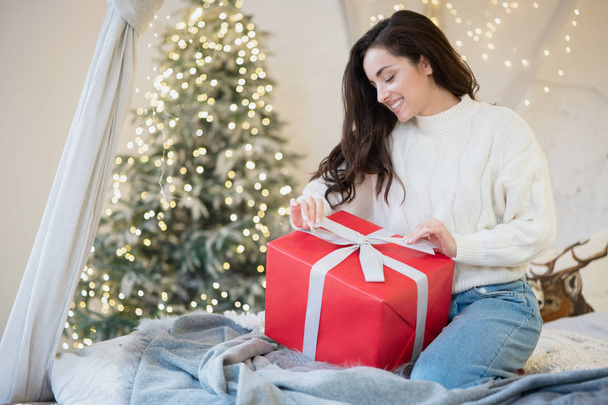 Young beautiful smiling woman wearing christmas sweater wrapping present in room decorated for celebrating the new year and christmas festive mood - 写真・画像