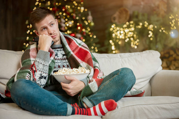 young handsome man wrapped in plaid watching TV eating popcorn on the sofa in room decorated for celebrating new year christmas all alone melancholic mood - Foto, Bild