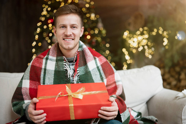 Young handsome man wearing warm sweater holding present sitting on the sofa in room decorated for celebrating the new year and christmas festive mood - Fotó, kép