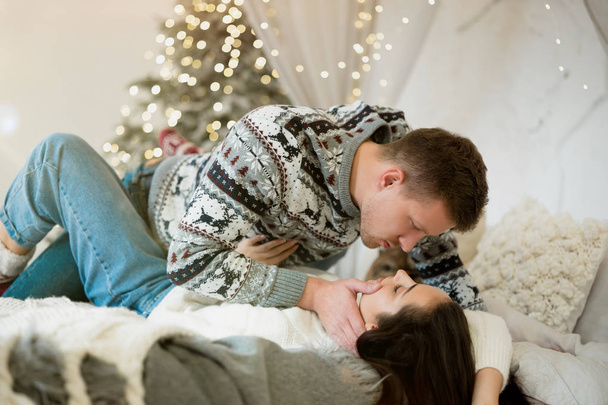 lovely couple beautiful woman and handsome man both wearing warm sweaters cuddling in bedroom decorated for celebrating the new year christmas festive mood - Photo, image