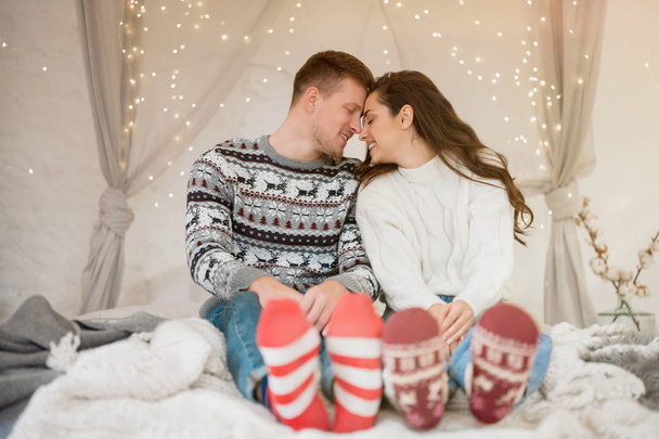 Young lovely couple beautiful wife and handsome husband both wearing warm sweaters and festive socks sitting in bedroom decorated for celebrating new year christmas tenderness - Fotografie, Obrázek