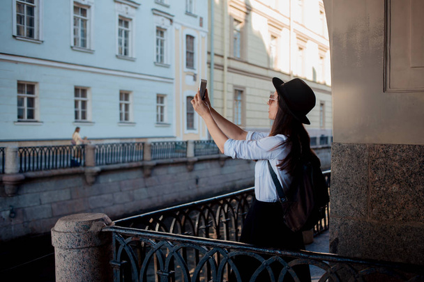 Tourist in the city takes a photo on smartphone. A young woman in a black hat and a white shirt, - Φωτογραφία, εικόνα