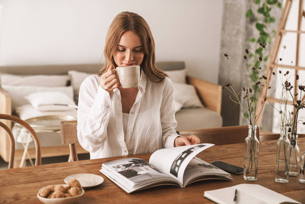 Woman sit indoors in office reading magazine drinking coffee. - Fotó, kép