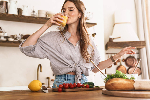 Pleased cute beautiful woman indoors at the kitchen cooking drinking juice. - Photo, Image