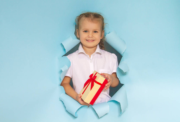 The head of a pretty girl peeps out of a hole in the paper. Girl holding a gift with a red bow.  Blue background. Festive Christmas composition. - Photo, Image
