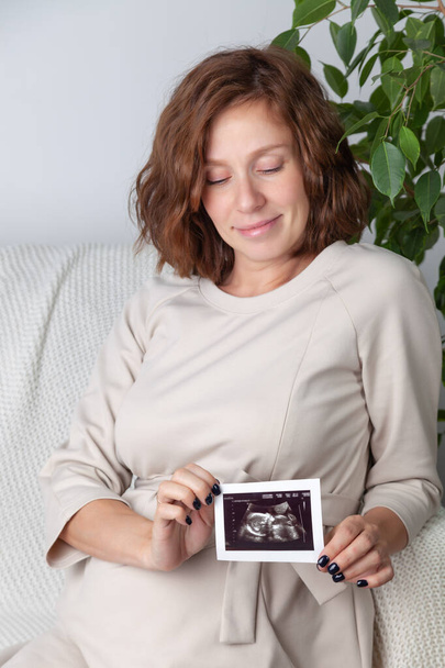 Pregnant woman holding ultrasound image. Concept of pregnancy, health care, gynecology, medicine. Young mother waiting of the baby. Close-up, copy space, indoors. - Photo, Image