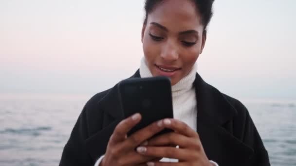 Дуже усміхнена афроамериканська дівчина радісно користується мобільним телефоном, що відпочиває біля моря.  - Кадри, відео