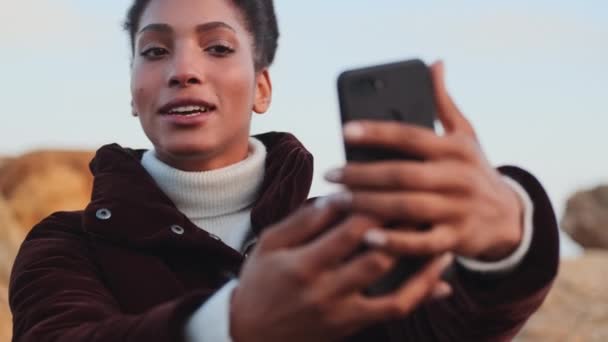 Kaunis hymyilevä Afrikkalainen Amerikan tyttö iloisesti puhuu aikana video chat matkapuhelin ulkona
 - Materiaali, video