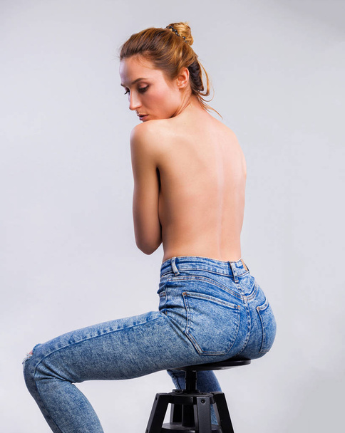Picture of beautiful topless woman in jeans sitting on chair covering breasts with hands posing in the studio on gray background - Фото, изображение