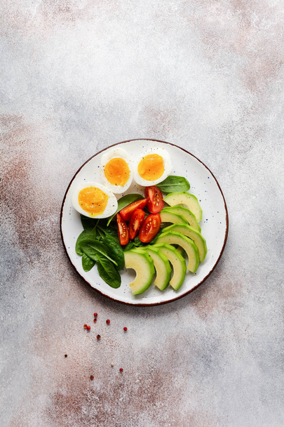 Healthy vegetarian salad breakfast. Spinach leaves, tomato, avocado and boiled egg on gray plate and gray concrete old background. Buddha bowl. Top view.  - Foto, Bild