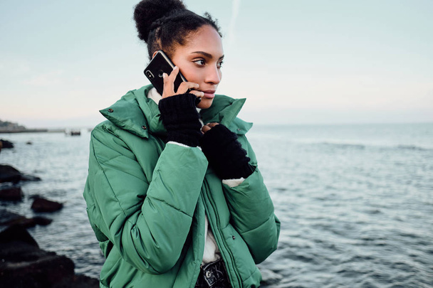 魅力的なアフリカ系アメリカ人の女の子の肖像-ダウンジャケットで携帯電話で海のそばを歩く - 写真・画像