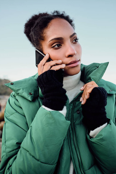 Pensativa chica afroamericana casual en chaqueta de plumón cuidadosamente mirando hacia otro lado hablando en el teléfono celular junto al mar
 - Foto, Imagen