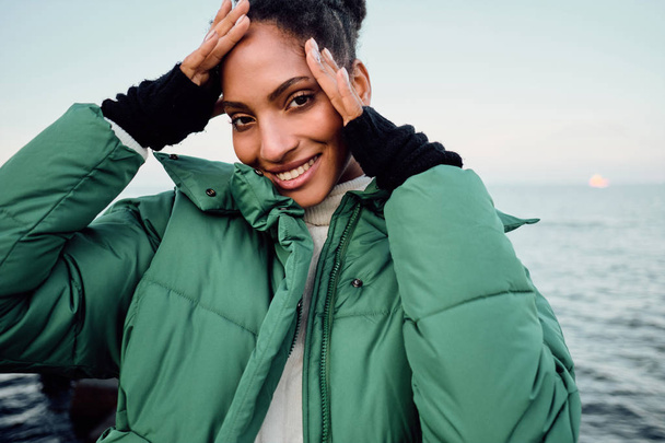 Portrait of pretty cute African American girl in stylish down jacket happily looking in camera by the sea - Photo, Image