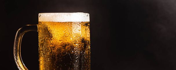 Studené pivo s pěnou v hrnku, na dřevěném stole a tmavém pozadí s prázdným místem pro logo nebo text. Stock Fotka hrnek studeného pěnového piva close-up. - Fotografie, Obrázek