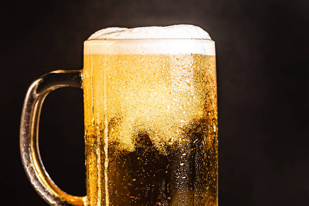 マグカップの泡と冷たいビール、木製のテーブルとロゴやテキストのための空白のスペースを持つ暗い背景。ストックフォト｜冷たい泡立ちビールのマグカップのクローズアップ. - 写真・画像
