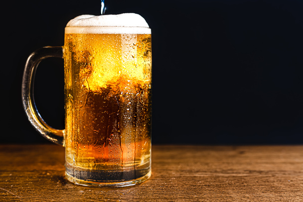 Κρύα μπύρα με αφρό σε κούπα, σε ξύλινο τραπέζι και σκούρο φόντο με κενό χώρο για λογότυπο ή κείμενο. Απόθεμα Photo κούπα κρύο αφρώδες μπύρα close-up. - Φωτογραφία, εικόνα