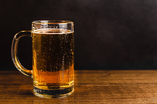 Cold beer with foam in a mug, on a wooden table and a dark background with blank space for a logo or text. Stock Photo mug of cold foamy beer close-up. - Foto, Bild