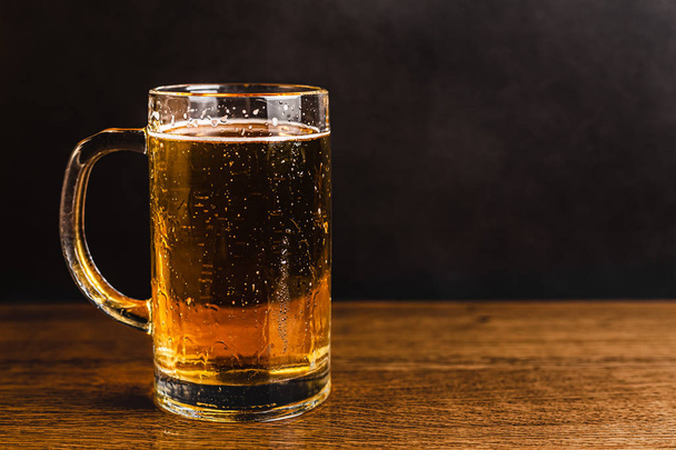 Cold beer with foam in a mug, on a wooden table and a dark background with blank space for a logo or text. Stock Photo mug of cold foamy beer close-up. - 写真・画像