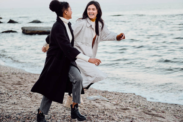 Две красивые стильные девушки в плащах счастливо бегут по морю
 - Фото, изображение