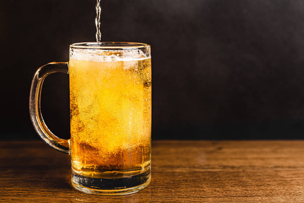 Cold beer with foam in a mug, on a wooden table and a dark background with blank space for a logo or text. Stock Photo mug of cold foamy beer close-up. - 写真・画像