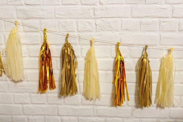Gold paper garlands on a white brick wall.  Tissue paper tassels garland Bunting happy birthday wedding party decor. Matte and glossy gold garland for party decoration - 写真・画像