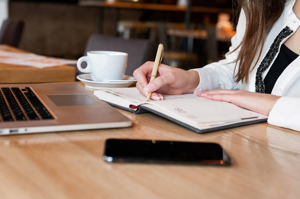 žena v bílé bundě pracuje ve svém notebooku dělat si poznámky ve svém plánovači při pití horké kávy v kavárně multitasking moderní businesswoman - Fotografie, Obrázek
