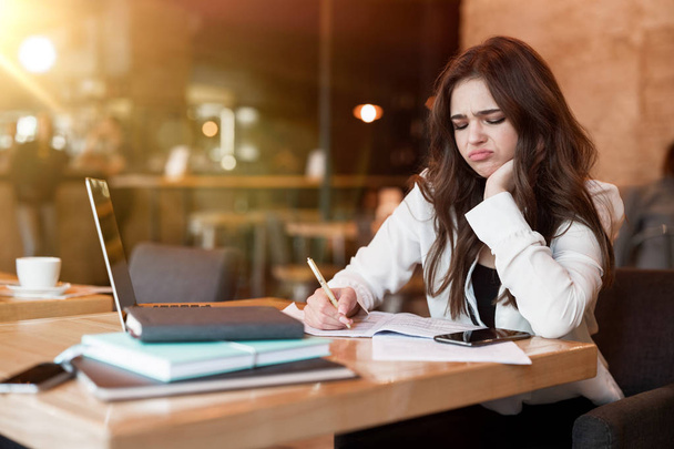 young woman in white jacket working in her laptop taking notes in her planner looking annoyed while drinking hot coffee in cafe multitasking modern businesswoman - Foto, Imagen