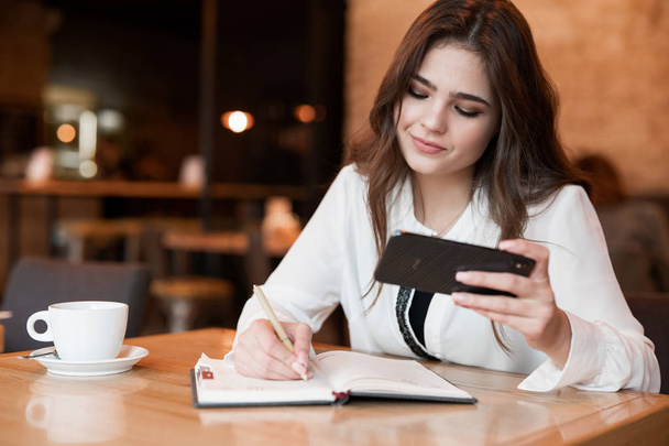 young beautiful woman taking notes to her planner using her smartphone drinking hot coffee in the cafe multitasking modern businesswoman - Photo, Image