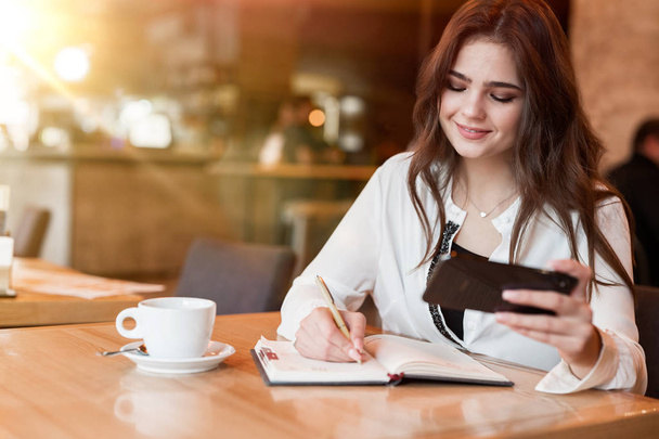 junge schöne Frau, die Notizen zu ihrem Planer macht, indem sie den Zeitplan ihres Smartphones benutzt und im Café heißen Kaffee trinkt, moderne Geschäftsfrau mit Multitasking - Foto, Bild