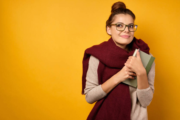 A brunette with glasses and a burgundy scarf, with a high bun clutching a book to herself and dreamily looks up. - Foto, imagen
