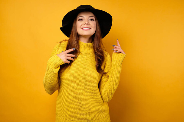 A beautiful brunette in a yellow sweater and black hat stands on a yellow background and looks into the frame. - Фото, изображение