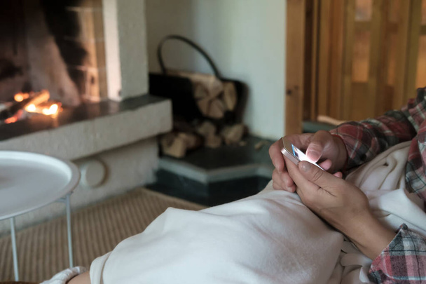 Femme avec un smartphone dans les mains, assise sur un canapé et recouverte d'un plaid, sur un fond flou d'une cheminée ouverte avec du bois de chauffage allumé
. - Photo, image